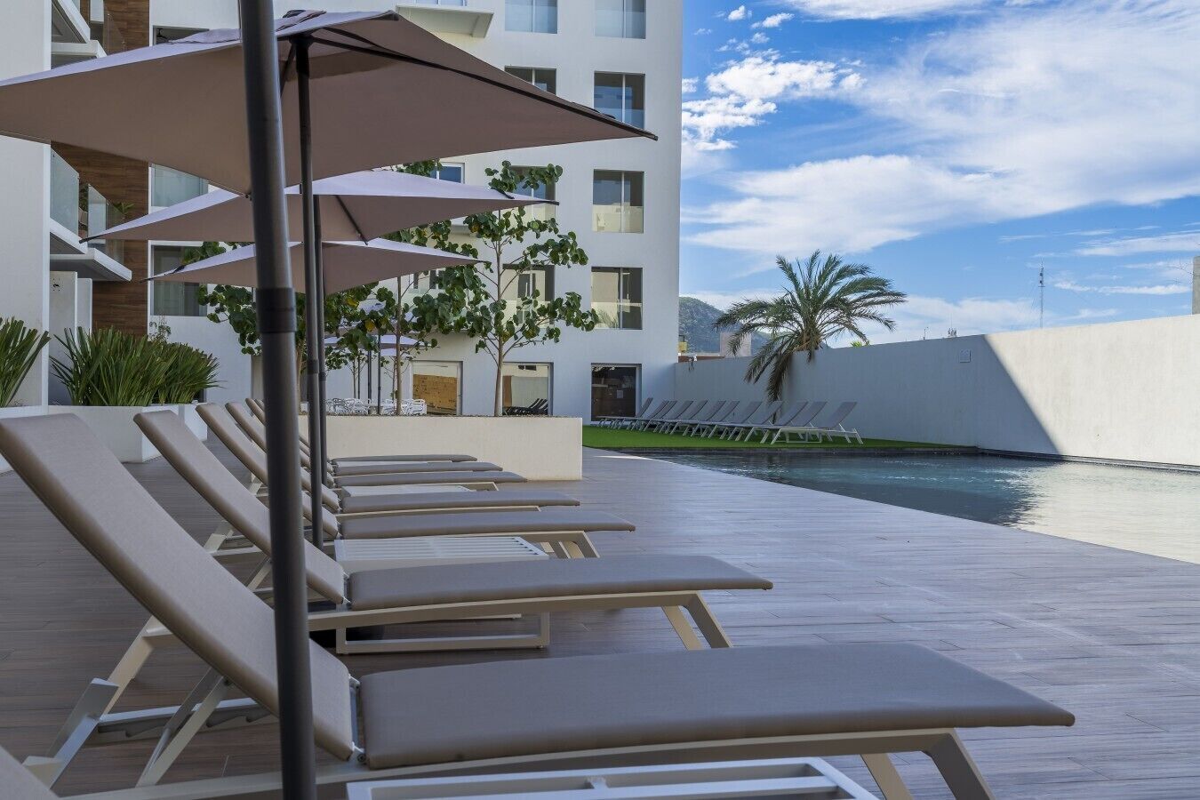 A poolside area with sun loungers and umbrellas, a clear sky, and a modern building in the background.