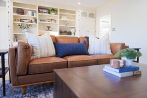 A brown leather sofa with cushions, a wooden coffee table, and a bookshelf filled with various items. - Huge Luxury Home | Game Room + Fenced Yard (Valdosta)