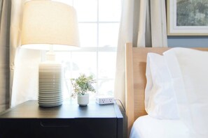 Ein ordentlich bezogenes Bett mit weißen Leinen, eine Nachttischlampe, ein kleiner Topfpflanze und ein Stapel weißer Teller auf einem Nachttisch.