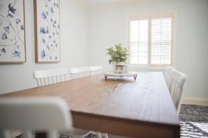 Ein Esszimmer mit Holztisch, weißen Stühlen, einer Topfpflanze und gerahmten Blumenbildern an der Wand.