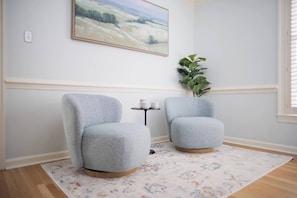 Ein Zimmer mit zwei grauen Sesseln, einem kleinen Tisch mit einer Tasse, einer Pflanze und einem Gemälde an der Wand.