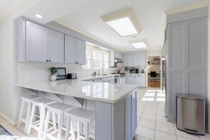 A modern kitchen with white cabinetry, a large island with bar stools, and a stainless steel trash can. - Huge Luxury Home | Game Room + Fenced Yard (Valdosta)