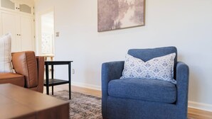 A living room with a blue armchair, a wooden coffee table, a patterned pillow, a black side table, and a painting on the wall. - Huge Luxury Home | Game Room + Fenced Yard (Valdosta)