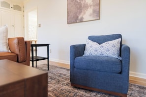 A living room with a blue armchair, a wooden coffee table, a patterned pillow, a black side table, and a painting on the wall.