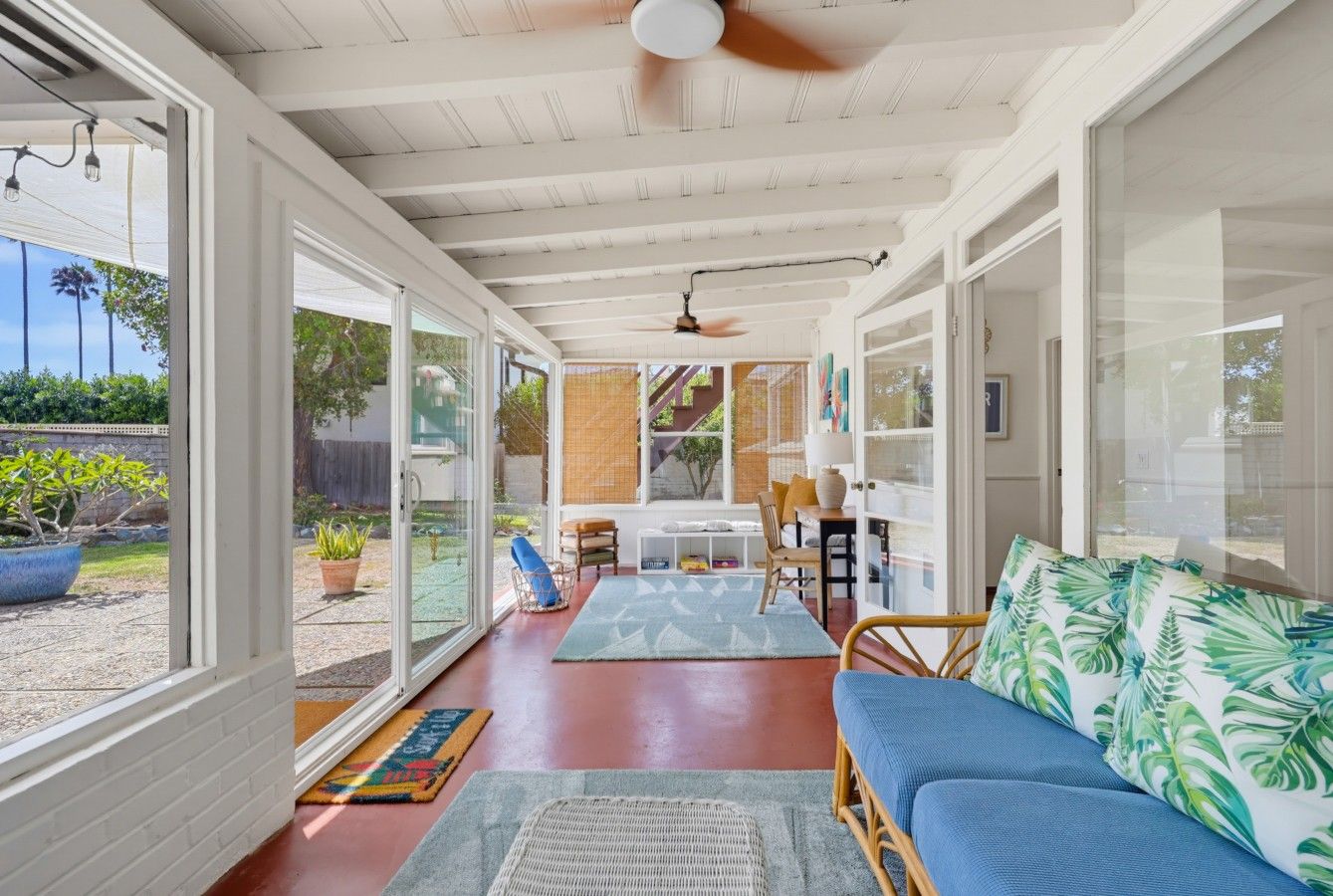 A sunlit room with a ceiling fan, a blue sofa with tropical print cushions, and a dining area with a table and chairs.