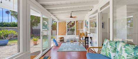 Una habitación soleada con ventilador de techo, un sofá azul con cojines estampados tropicales y un comedor con mesa y sillas.