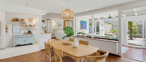 Un espace repas avec une table et des chaises en bois, un luminaire suspendu et une vue sur l’extérieur grâce à de grandes fenêtres.