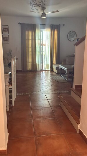 A spacious living area with a tiled floor, a ceiling fan, and a view through a window with curtains.