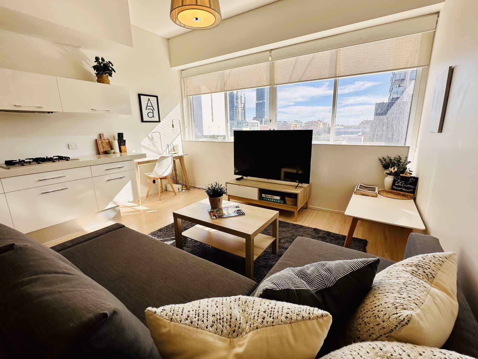 A modern living room with a sofa, coffee table, and a TV. There is a kitchen area with cabinets and a stove.