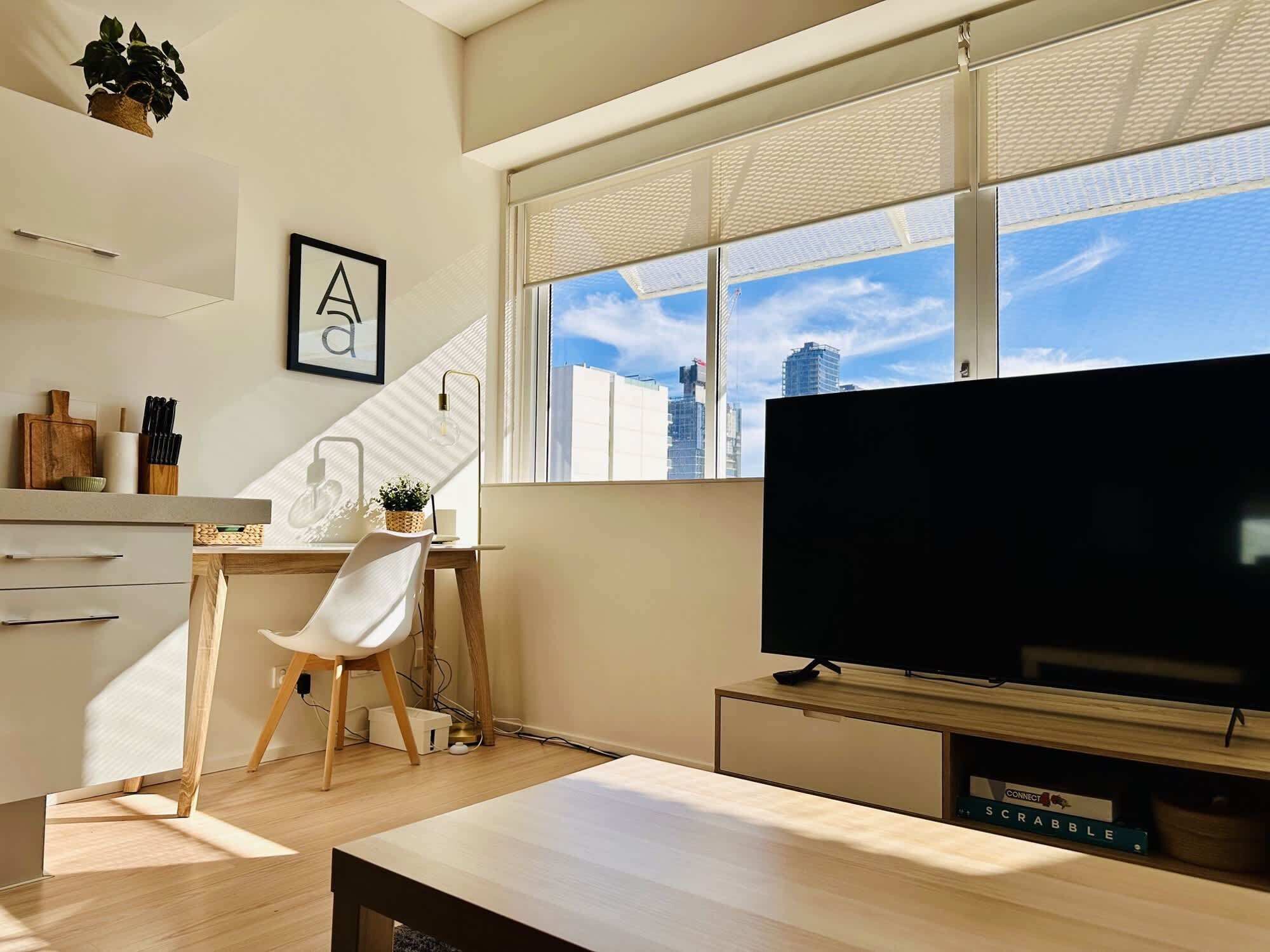 Un espacio habitable moderno con un gran ventanal, televisor de pantalla plana, escritorio con silla y zona de cocina.