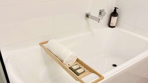 A white bathtub with a wooden tray holding a rolled towel and a bottle of shampoo. - Stylish 1-Bed Apartment in Adelaide CBD (Adelaide)