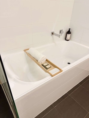 A white bathtub with a wooden tray holding a rolled towel and a bottle of shampoo. - Stylish 1-Bed Apartment in Adelaide CBD (Adelaide)