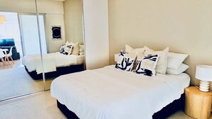 A bedroom with a large mirror, a bed with white bedding and patterned pillows, a bedside table with a lamp, and a view into another room with a desk and chair. - Stylish 1-Bed Apartment in Adelaide CBD (Adelaide)