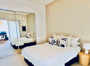 A bedroom with a large mirror, a bed with white bedding and patterned pillows, a bedside table with a lamp, and a view into another room with a desk and chair. - Stylish 1-Bed Apartment in Adelaide CBD (Adelaide)
