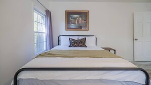 A single bed with a headboard, a bedside table, a window with curtains, and a framed picture on the wall. - Sleeps 18 Riverfront Getaway with Game Room & Deck (Valdosta)