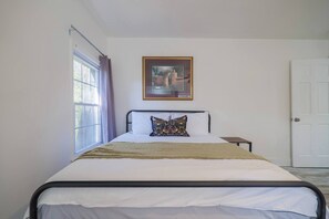 Ein Einzelbett mit Kopfteil, ein Nachttisch, ein Fenster mit Vorhängen und ein gerahmtes Bild an der Wand.