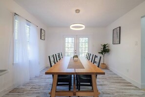 A dining room with a long wooden table, black chairs, white curtains, and a hanging light fixture. - Sleeps 18 Riverfront Getaway with Game Room & Deck (Valdosta)