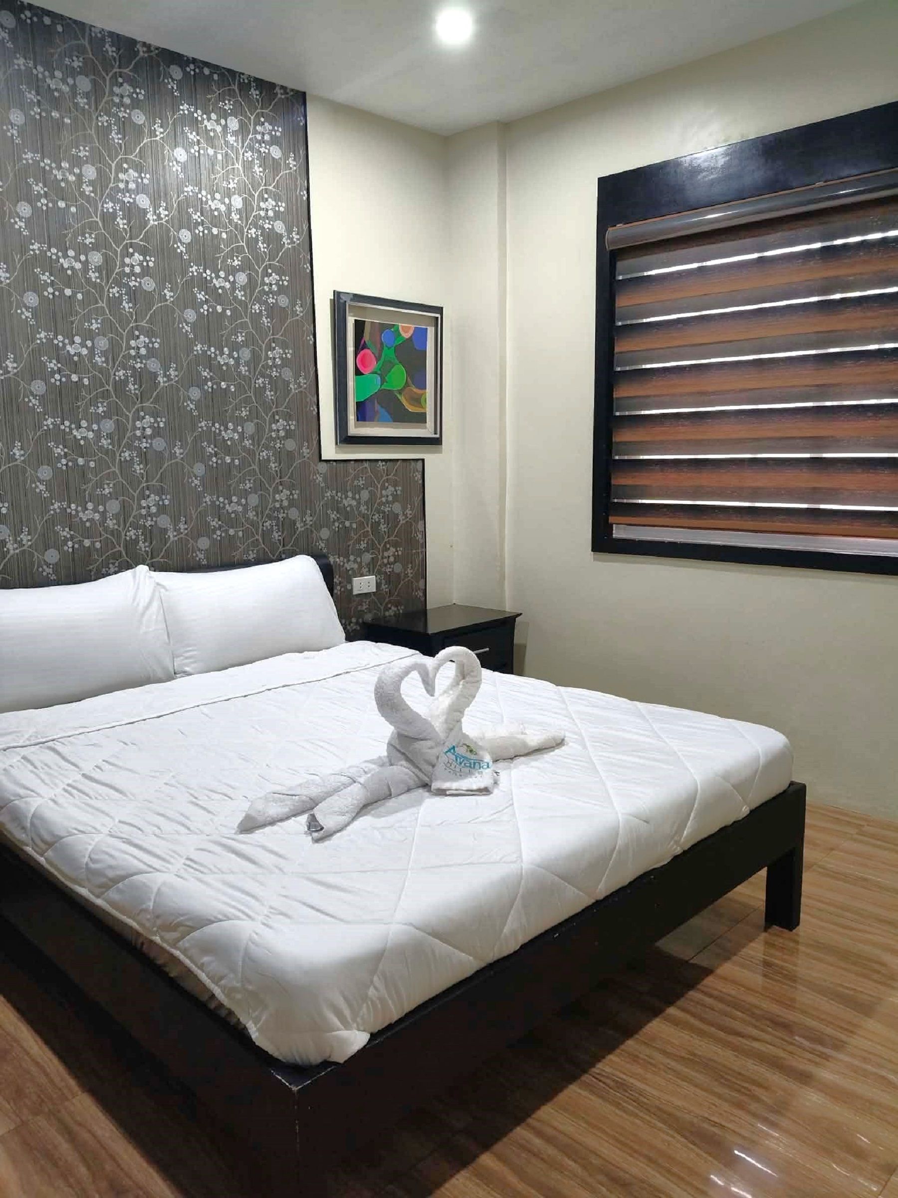 A hotel room with a bed, a nightstand, a framed picture, and a window with blinds.