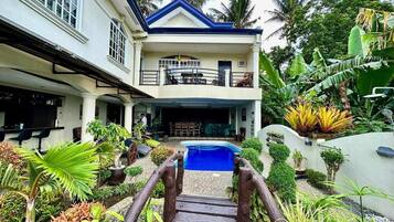 A two-story house with a pool, surrounded by tropical plants and palm trees.