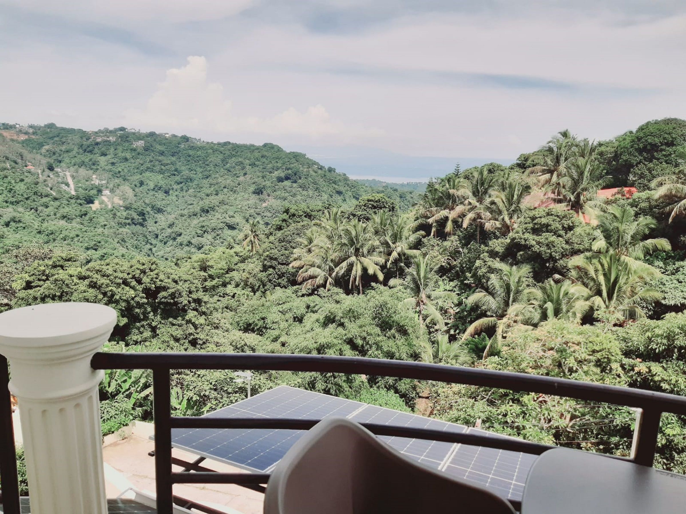 A balcony with solar panels, a white column, and a view of a lush green landscape with palm trees.