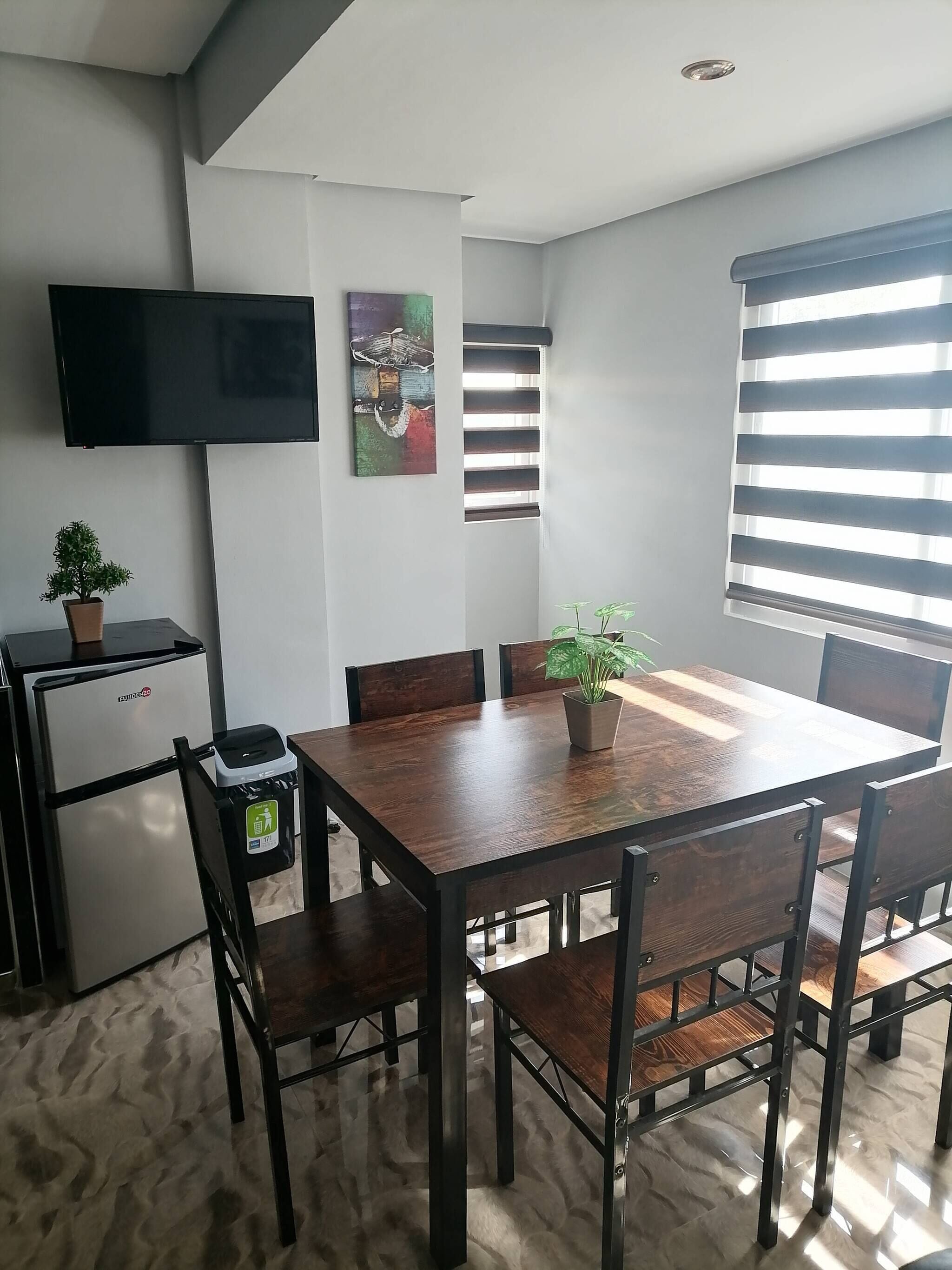 A dining area with a wooden table and chairs, a television mounted on the wall, a small refrigerator, and a potted plant.