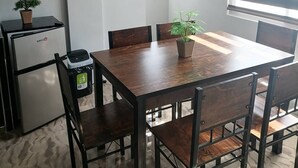 A dining area with a wooden table and chairs, a television mounted on the wall, a small refrigerator, and a potted plant.