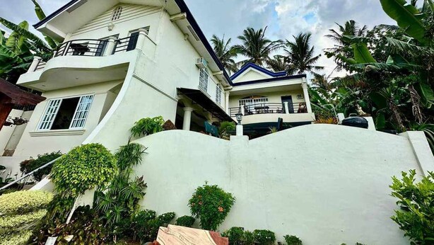A two-story building with a balcony on the upper floor, surrounded by lush greenery and palm trees.
