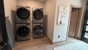 A laundry room with two stacked front-loading washing machines, a wooden floor, a door leading to another room, and a wall-mounted artwork. - My Place in Powder House Pass, Paved Roads, AC, Hot Tub! (Lead)