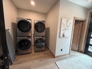 A laundry room with two stacked front-loading washing machines, a wooden floor, a door leading to another room, and a wall-mounted artwork. - My Place in Powder House Pass, Paved Roads, AC, Hot Tub! (Lead)