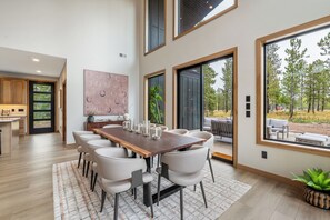 Un vaste espace dédié à la salle à manger, avec une longue table en bois et des chaises blanches, de grandes fenêtres offrant une vue sur les arbres, et une cuisine moderne en arrière-plan.