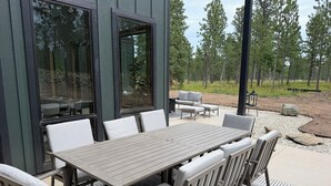 An outdoor patio with a wooden table and chairs, a sofa, and a large window reflecting trees. - My Place in Powder House Pass, Paved Roads, AC, Hot Tub! (Lead)