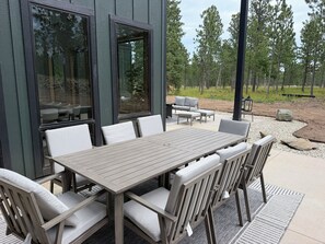 An outdoor patio with a wooden table and chairs, a sofa, and a large window reflecting trees. - My Place in Powder House Pass, Paved Roads, AC, Hot Tub! (Lead)