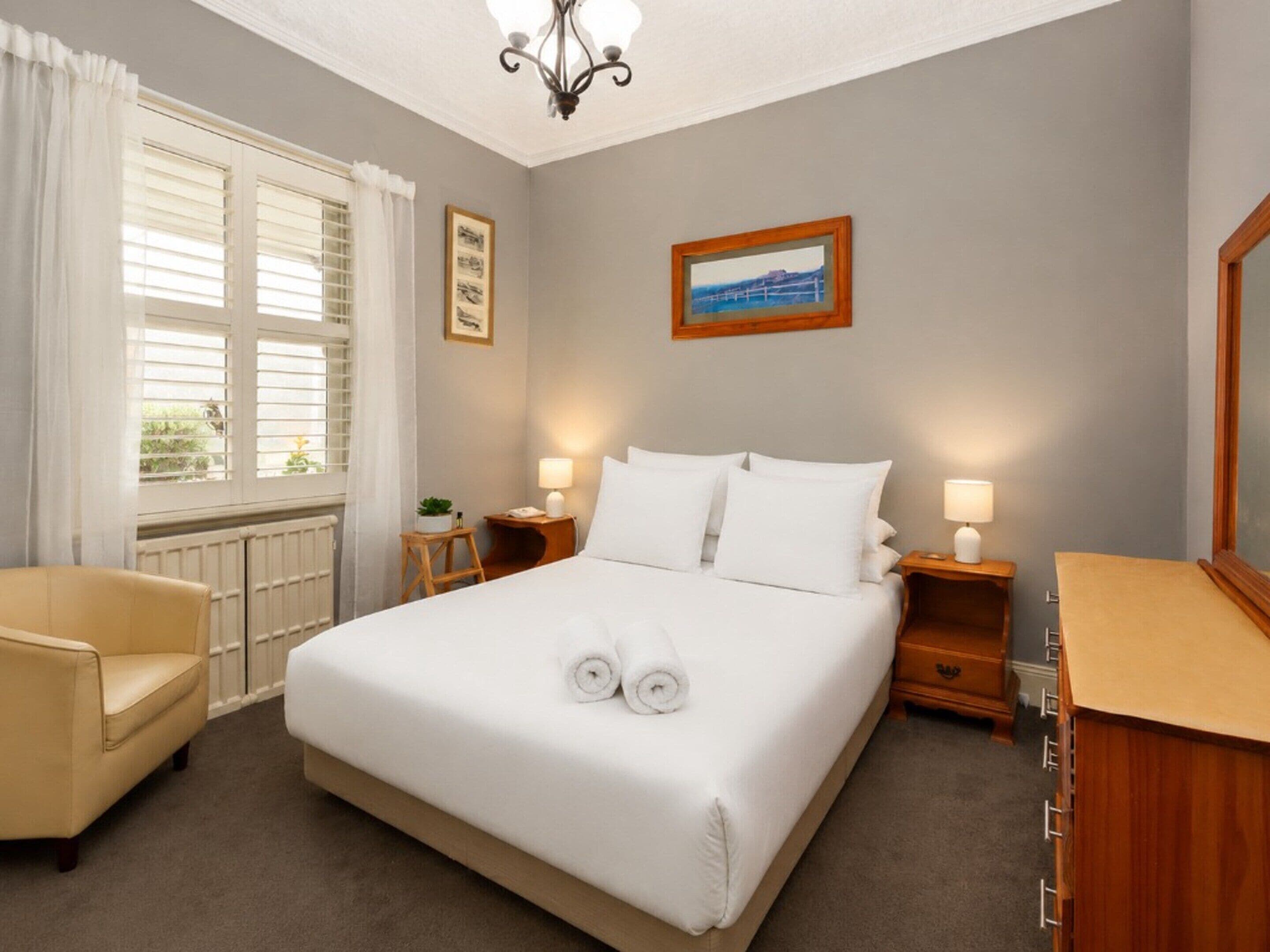 A hotel room with a large bed, two bedside tables with lamps, a chair, a window with curtains, and a framed picture on the wall.