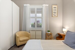 Un dormitori amb un llit, una cadira, un radiador, una finestra amb cortines, una taula petita amb una planta i una làmpada.