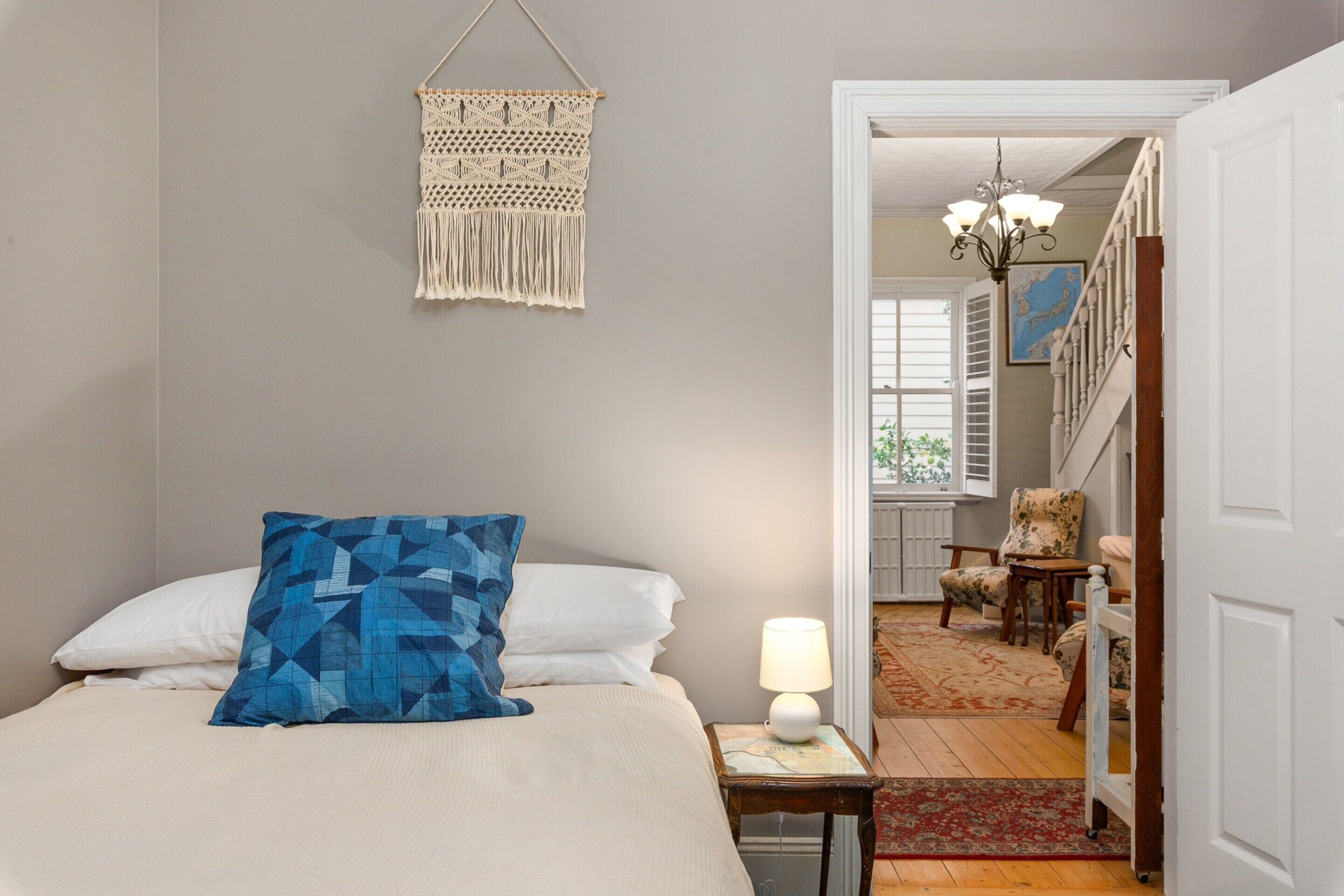 Un dormitorio con una cama, una decoración de pared de macramé colgante, una lámpara en una mesita de noche y vistas a una escalera que lleva a otra habitación.