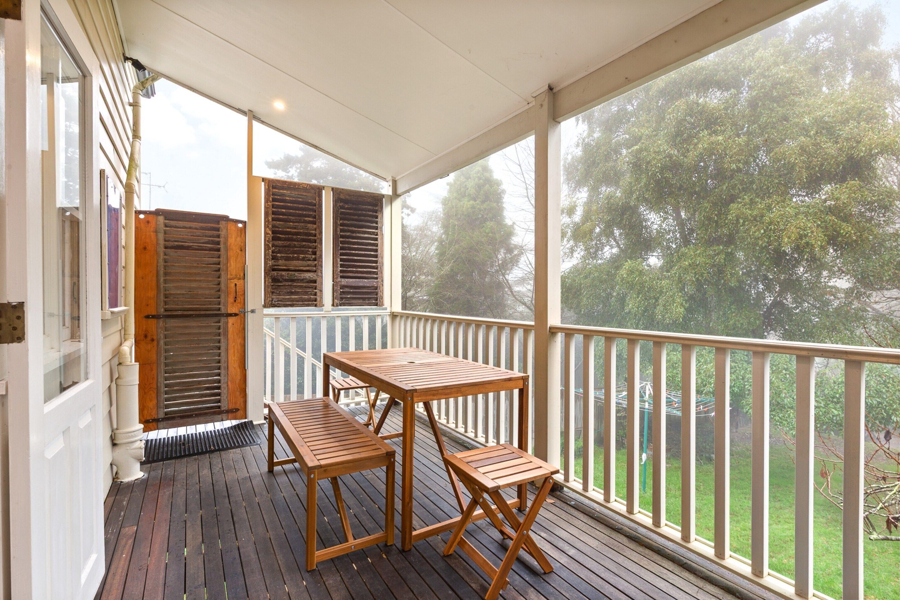 A covered outdoor area with a wooden table and benches, a view of trees through the railing, and a door with shutters.
