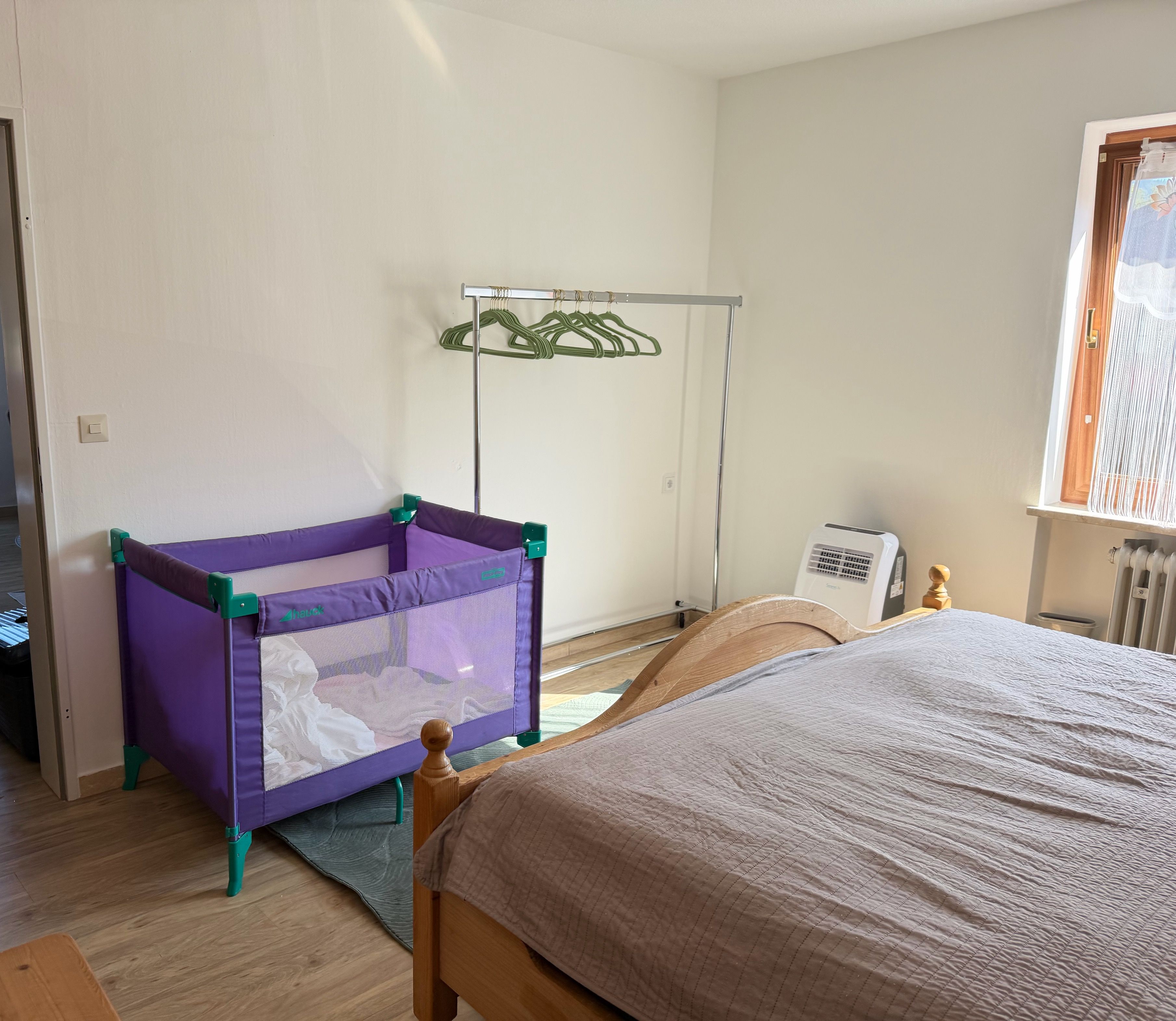 A bedroom with a bed, a purple baby playpen, a radiator, and a window.