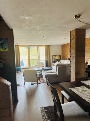 A spacious living room with a wooden floor, a sofa, a dining table, and a view of the outdoors through large windows. - 4BR, 4 full bathroom condo, with community pool. Near Lake Michigan. (Cedar)