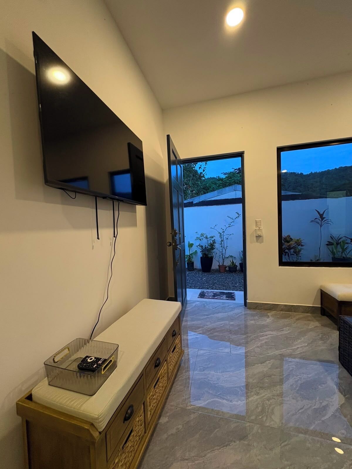 A modern interior with a flat-screen TV mounted on the wall, a bench with a remote control, and a view of a garden through an open door.