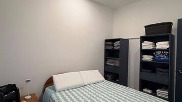 Een slaapkamer met een bed, nachtkastje, lamp en ingebouwde planken met opgevouwen kleding.