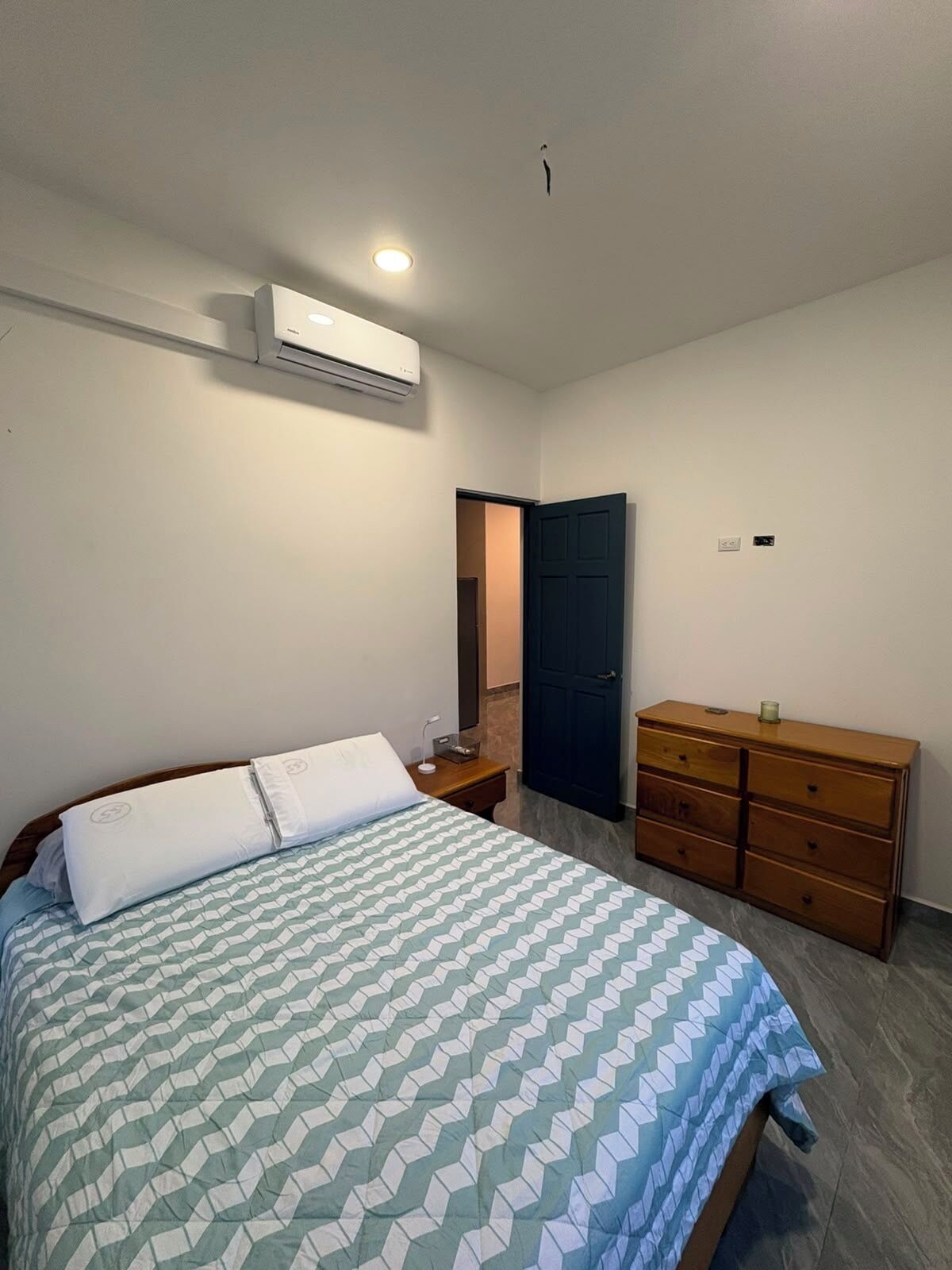 A bedroom with a bed, a dresser, an air conditioning unit, and a door leading to another room.