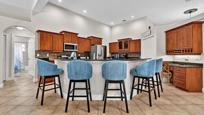A modern kitchen with wooden cabinets, a central island with bar stools, and a view to the adjacent bathroom. - Sea Your Escape | Pool, Game Room & Lanai | Palm Coast Near Flagler Beach (Palm Coast)