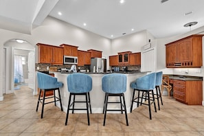 A modern kitchen with wooden cabinets, a central island with bar stools, and a view to the adjacent bathroom. - Sea Your Escape | Pool, Game Room & Lanai | Palm Coast Near Flagler Beach (Palm Coast)