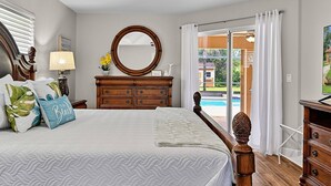 A bedroom with a bed, a dresser, a mirror, and a view of a pool. - Sea Your Escape | Pool, Game Room & Lanai | Palm Coast Near Flagler Beach (Palm Coast)