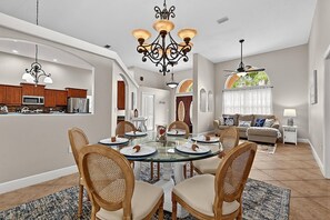 A dining area with a glass table surrounded by wicker chairs, a chandelier, and a view of the kitchen and living room. - Sea Your Escape | Pool, Game Room & Lanai | Palm Coast Near Flagler Beach (Palm Coast)