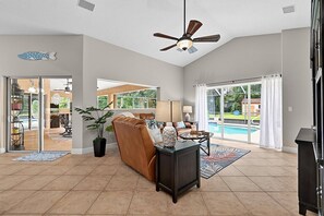 A spacious living area with a ceiling fan, a pool visible through large sliding glass doors, and a dining area with a table and chairs. - Sea Your Escape | Pool, Game Room & Lanai | Palm Coast Near Flagler Beach (Palm Coast)