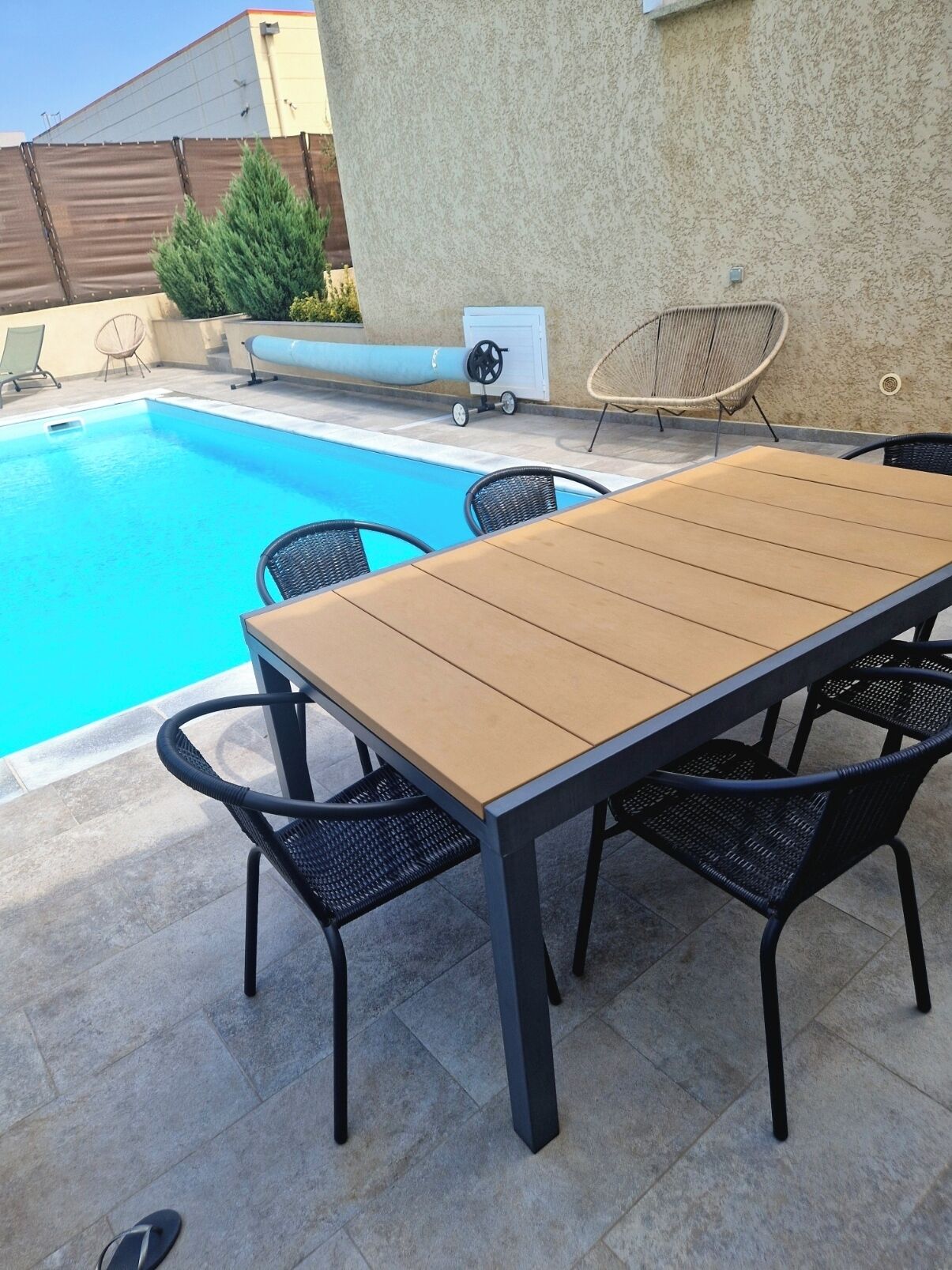 An outdoor pool area with a wooden table and chairs, a lounge chair, and a small water feature.