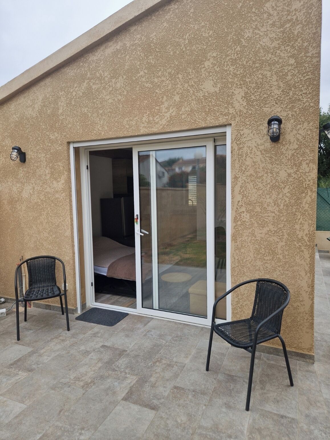 A building with a sliding glass door leading to a patio area with two chairs and a bed visible inside.