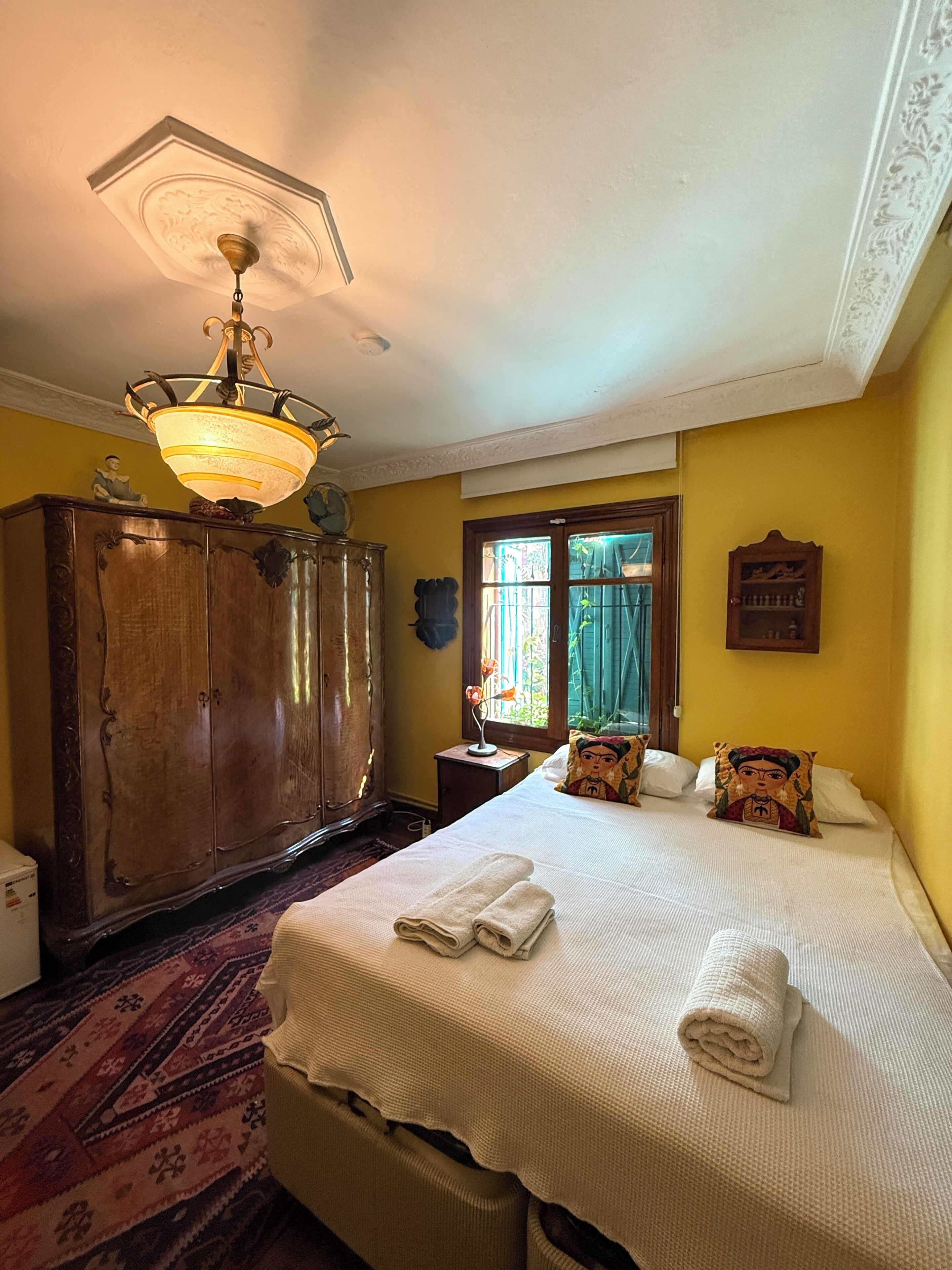 A room with a bed, a wooden wardrobe, a chandelier, and a window with curtains.