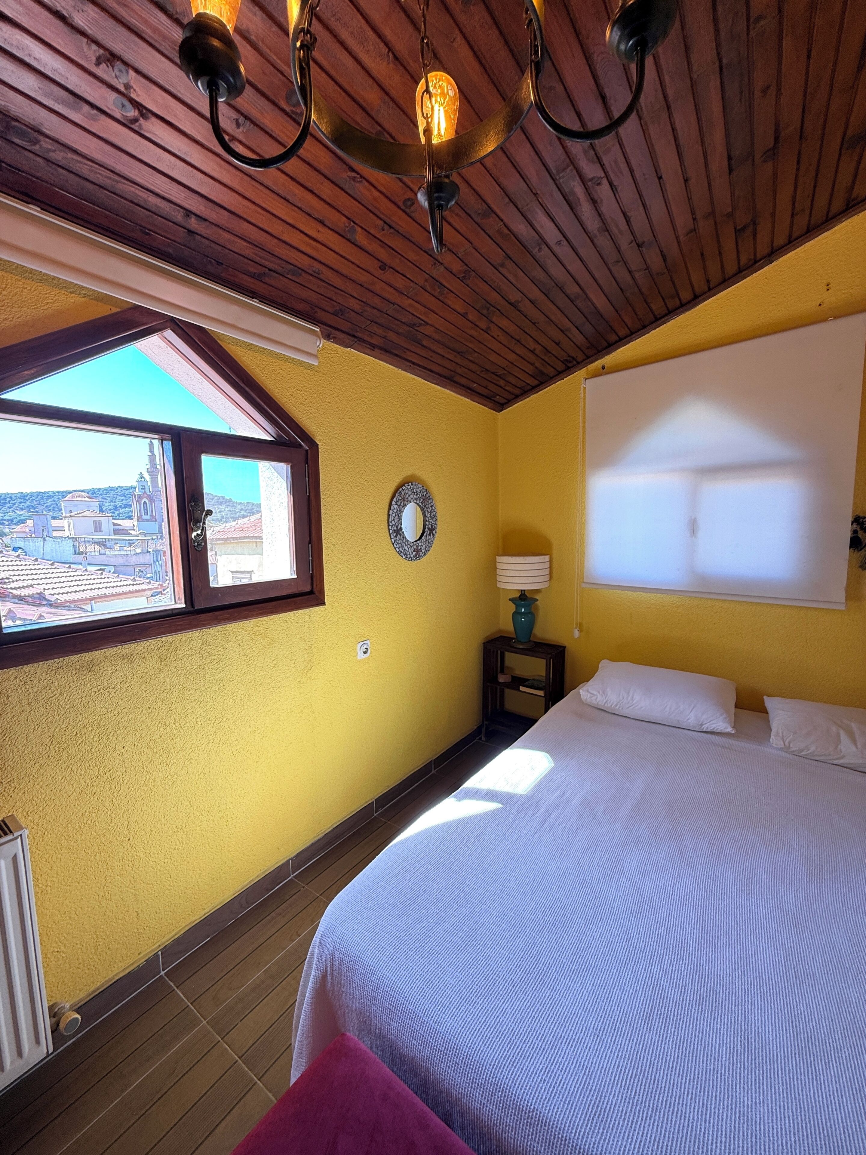 Un dormitorio con techo de madera, una cama, una ventana con vistas, un reloj de pared y una lámpara de noche.